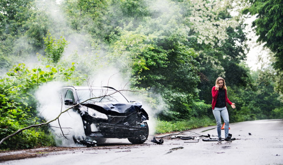 A young woman with smartphone by the damaged car after a car accident, making a phone call.