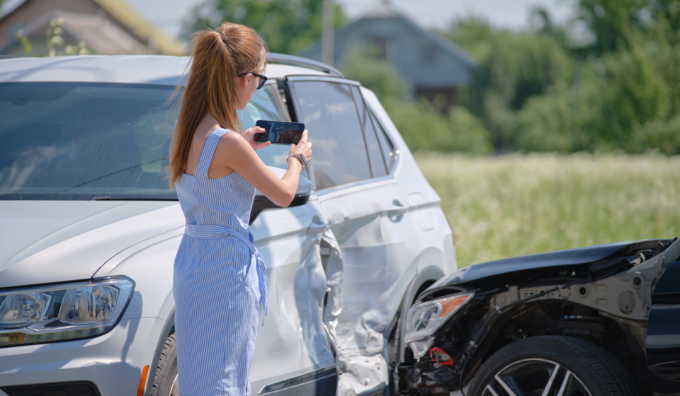 Stressed woman driver taking photo on mobile phone camera after vehicle collision on street side for emergency service after car accident. Road safety and insurance concept